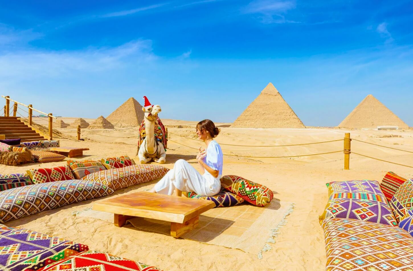A woman enjoys sitting by the pyramids with a decorated camel in Christmas attire and colorful cushions around her, perfectly capturing the spirit of Egypt Christmas holiday packages.